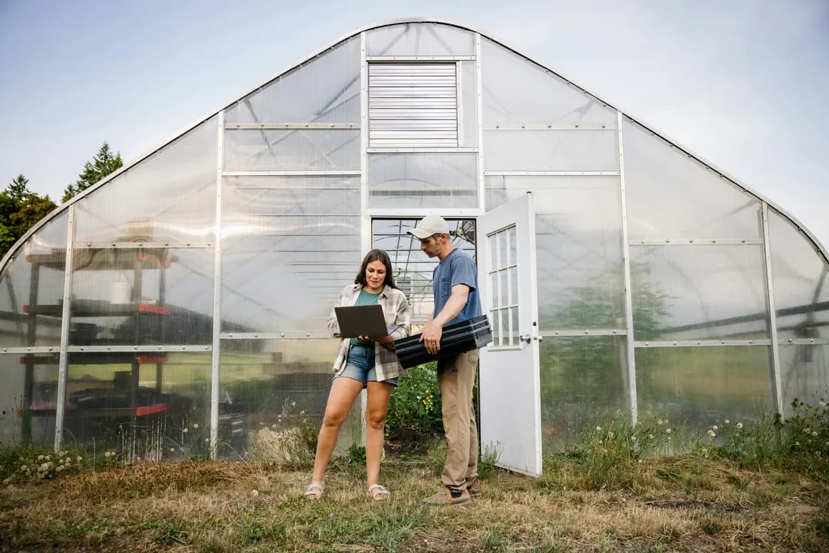 Couple d'agriculteurs devant une serre, ordinateur en main, préparant une demande d'aide à l'investissement